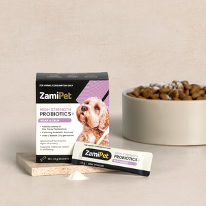 Styled image showing a box of ZamiPet High Strength Probiotics+ Relax & Calm, a probiotic sachet and small pile of probiotic powder placed in front. Ceramic bowl filled with dry dog food in background. 