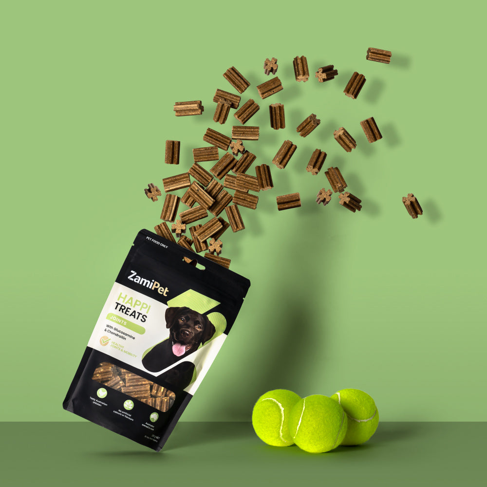 A pouch of ZamiPet HappiTreats Joints with treats exploding out the top in an arc. The background is light green and the pouch is sitting next to a small pile of yellow tennis balls